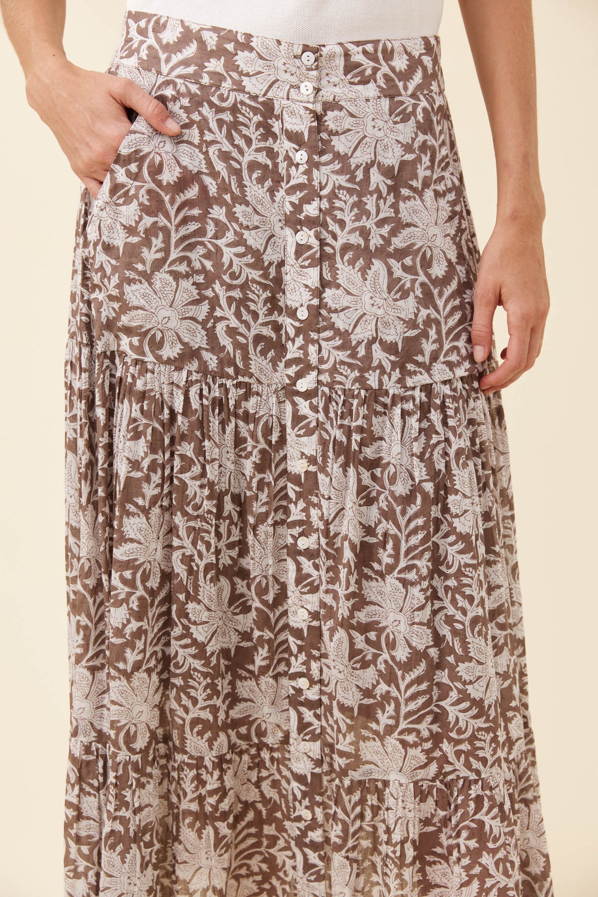 A close-up of a person wearing a brown and beige floral print skirt with a button-front waistband and pleated tiers, against a plain light beige background.