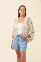 A woman modeling a cream-colored crochet cardigan over a white tank top and light blue denim shorts, standing against a solid light beige background.