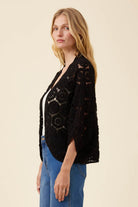 A woman wearing a black open-front crochet cardigan with three-quarter sleeves over a white top and blue jeans, photographed against a plain beige studio background.