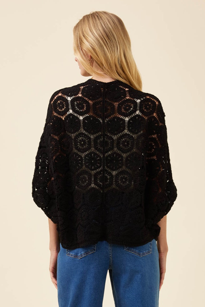 A woman viewed from the back wearing a black open-knit crochet cardigan with a geometric hexagonal pattern, paired with blue jeans, against a solid light beige background.
