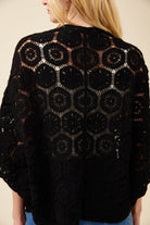 A woman shown from the back wearing a black crocheted openwork cardigan or poncho with a repeating circular lace pattern, against a plain light beige studio background.
