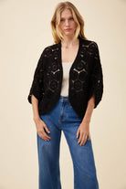 A woman modeling a black open-front crochet cardigan with three-quarter sleeves, worn over a white ribbed tank top and blue jeans, against a solid beige background.
