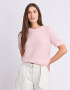 A woman modeling a light pink short-sleeve knit sweater with a crew neck, paired with off-white trousers, against a plain light gray studio background.