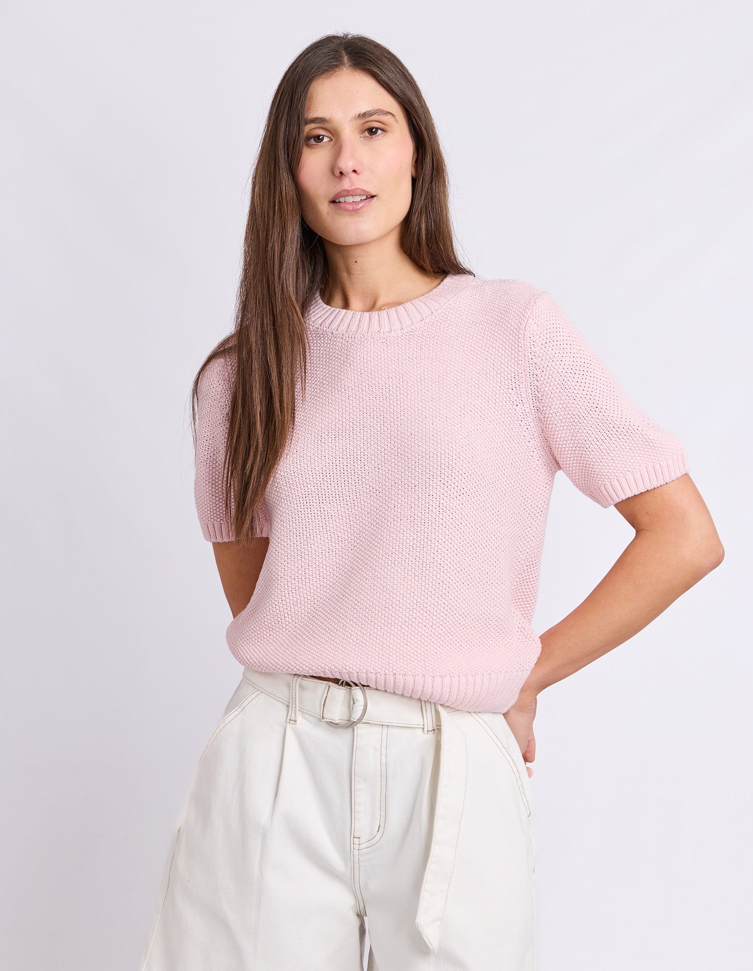 A woman modeling a light pink short-sleeve knit sweater with a crew neck, paired with off-white trousers, against a plain light gray studio background.