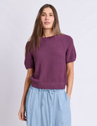 A woman modeling a short-sleeve knit sweater in a muted plum purple color, paired with light blue drawstring pants, against a plain light gray studio background.
