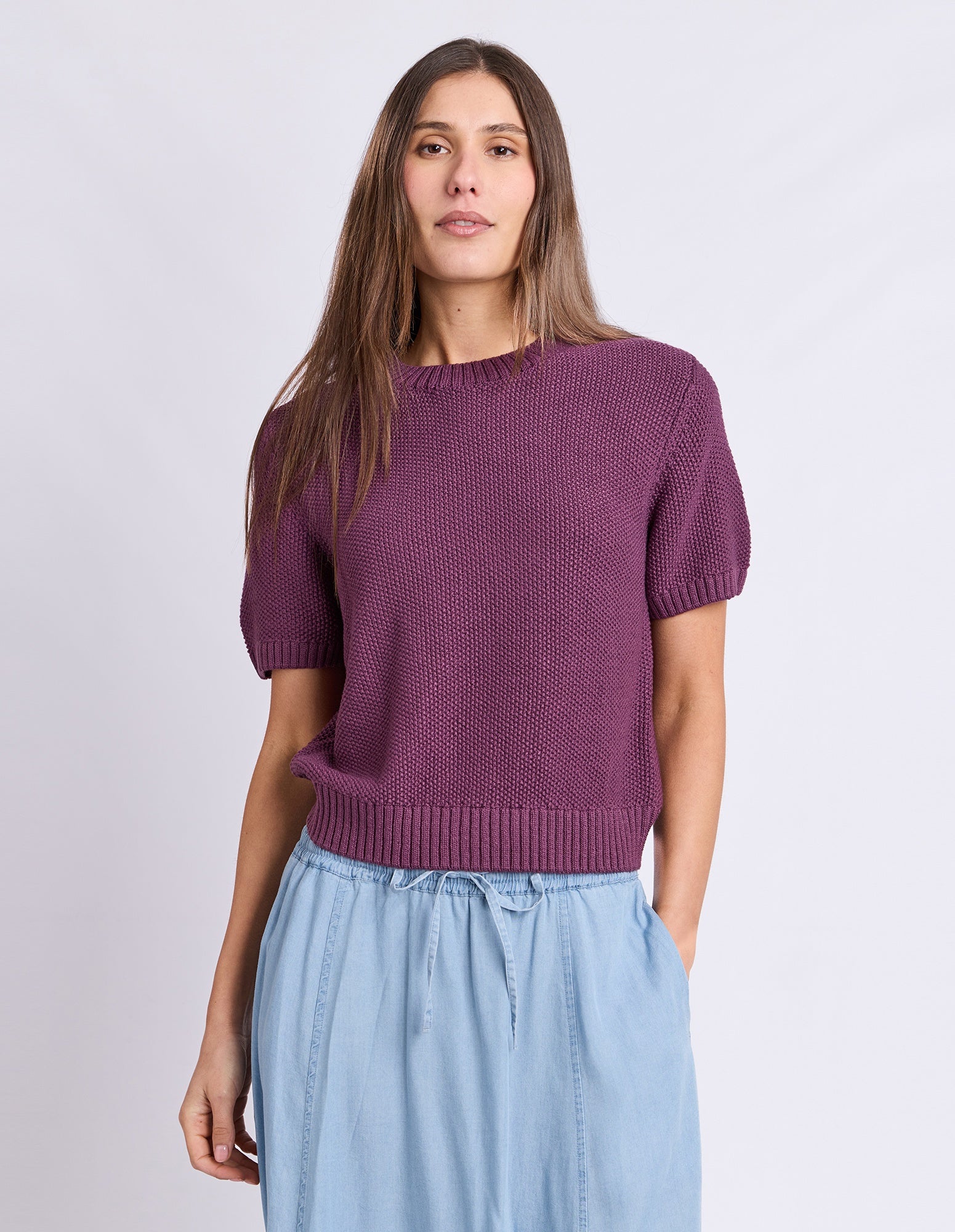 A woman modeling a short-sleeve knit sweater in a muted plum purple color, paired with light blue drawstring pants, against a plain light gray studio background.