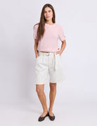 A woman modeling a light pink short-sleeve knit top and off-white pleated shorts with a tie belt, paired with dark brown flats, against a light gray studio background.