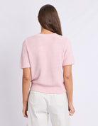 A woman viewed from the back wearing a light pink short-sleeve knit sweater and off-white trousers against a plain light gray studio background.