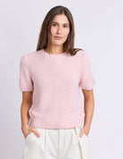 A woman wearing a light pink short-sleeve knit sweater and off-white trousers, standing against a plain light gray studio background.