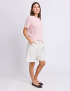 A woman modeling a light pink short-sleeve knit sweater and off-white shorts against a plain light gray studio background.