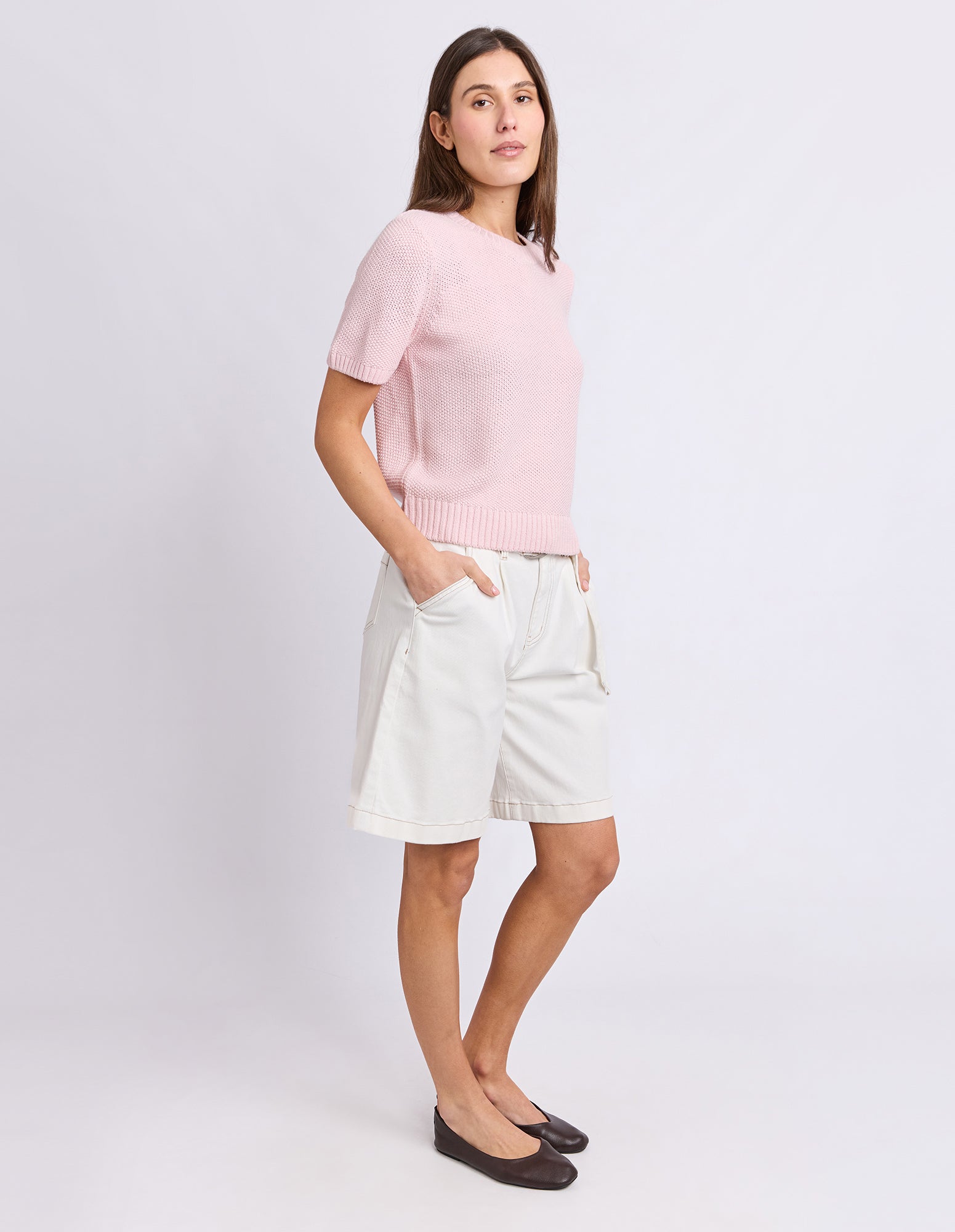 A woman modeling a light pink short-sleeve knit sweater and off-white shorts against a plain light gray studio background.