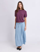 A woman modeling a short-sleeve purple knit top and a light blue midi skirt with a drawstring waist, paired with black flats, against a light gray studio background.