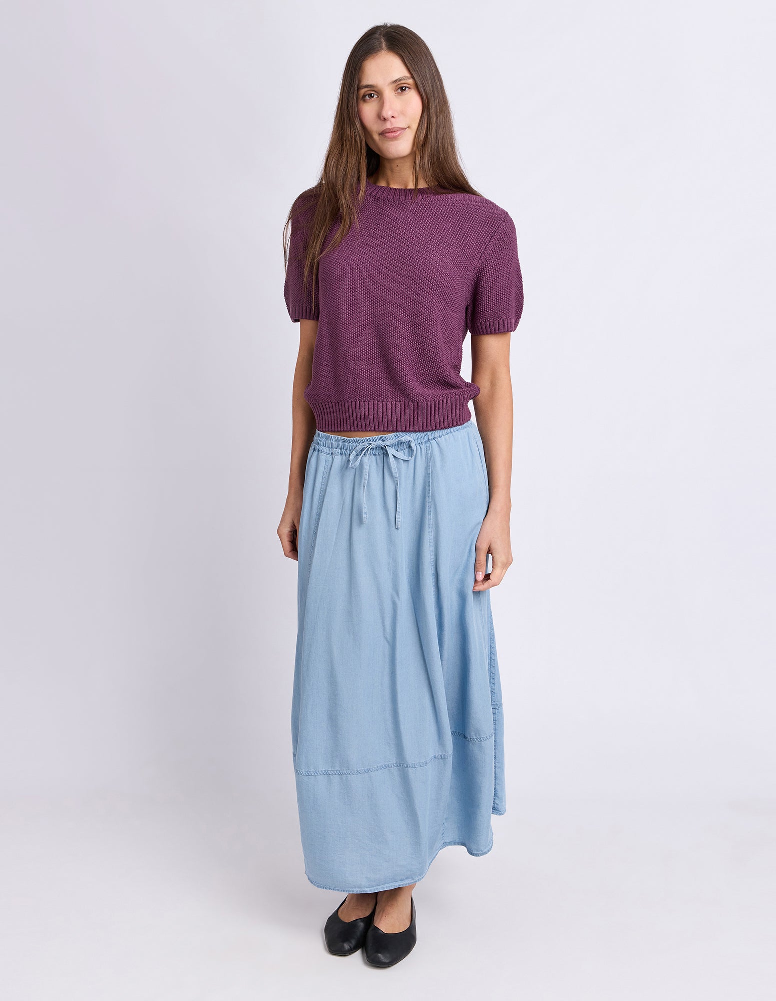A woman modeling a short-sleeve purple knit top and a light blue midi skirt with a drawstring waist, paired with black flats, against a light gray studio background.