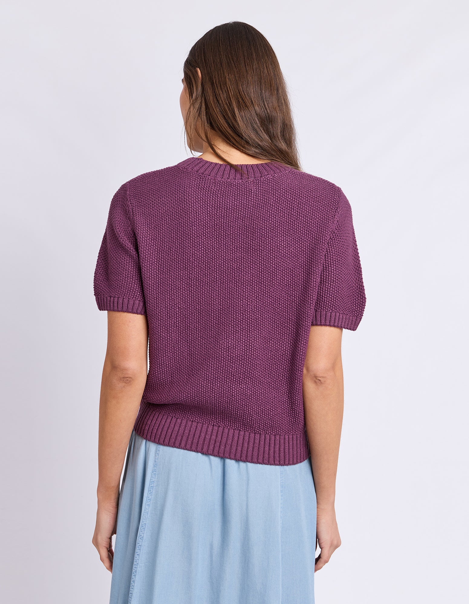 A woman shown from the back wearing a short-sleeve textured knit sweater in a muted plum purple, paired with a light blue skirt, against a plain light gray studio background.