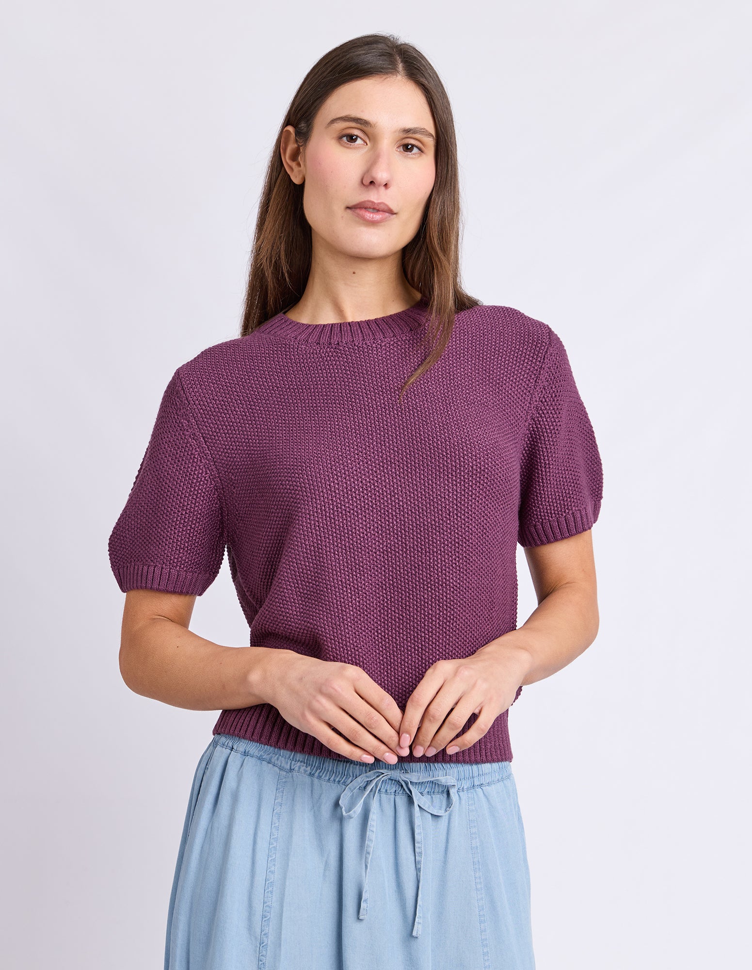 A woman wearing a short-sleeve textured knit top in a muted plum purple, paired with light blue drawstring pants, against a plain light gray studio background.