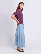 A woman modeling a short-sleeve purple knit top and a light blue wide-leg skirt, paired with black flats, against a plain light gray studio background.