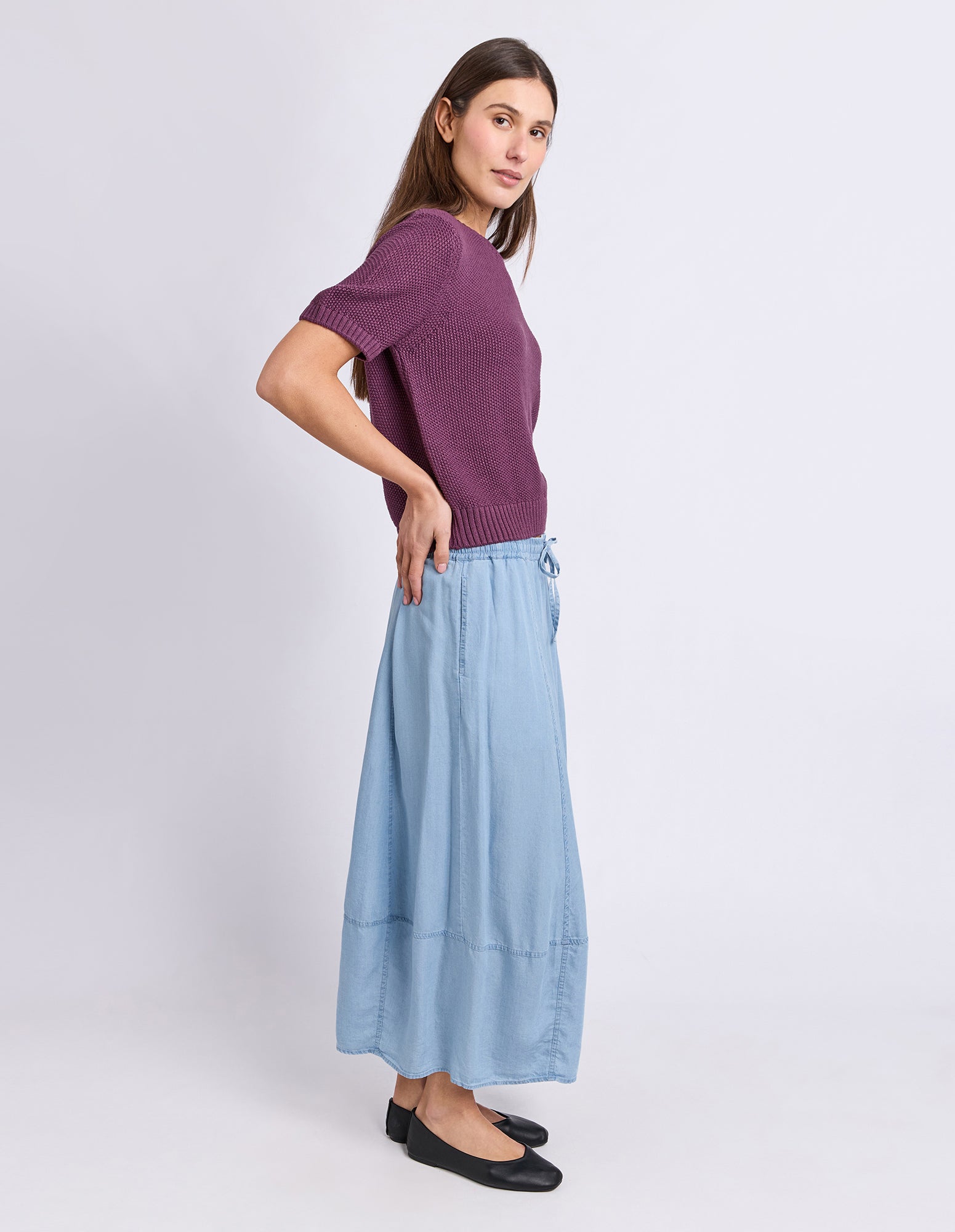 A woman modeling a short-sleeve purple knit top and a light blue wide-leg skirt, paired with black flats, against a plain light gray studio background.