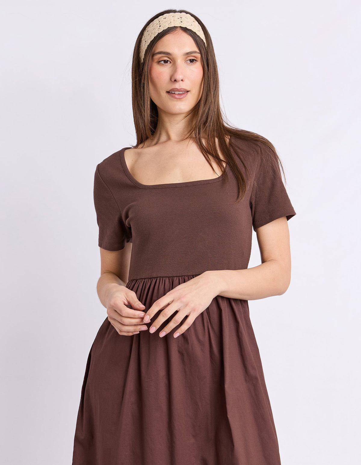 A woman modeling a brown short-sleeve dress with a square neckline and a gathered skirt, paired with a beige patterned headband, against a light gray studio background.