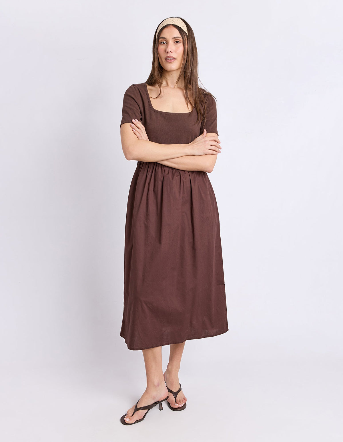 A woman modeling a brown short-sleeve midi dress with a square neckline and gathered waist, paired with black thong sandals and a cream headband, against a light gray studio background.