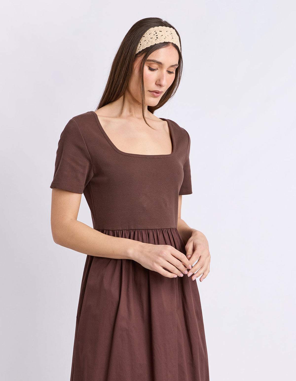A woman wearing a brown short-sleeve dress with a square neckline and a pleated waist, paired with a beige patterned headband, photographed against a light gray studio background.