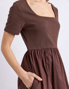 A close-up of a model wearing a brown short-sleeve dress with a square neckline and a gathered waist.