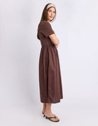 A woman modeling a brown short-sleeve midi dress with a gathered waist, paired with black heeled sandals and a beige patterned headband, standing against a light gray studio background.
