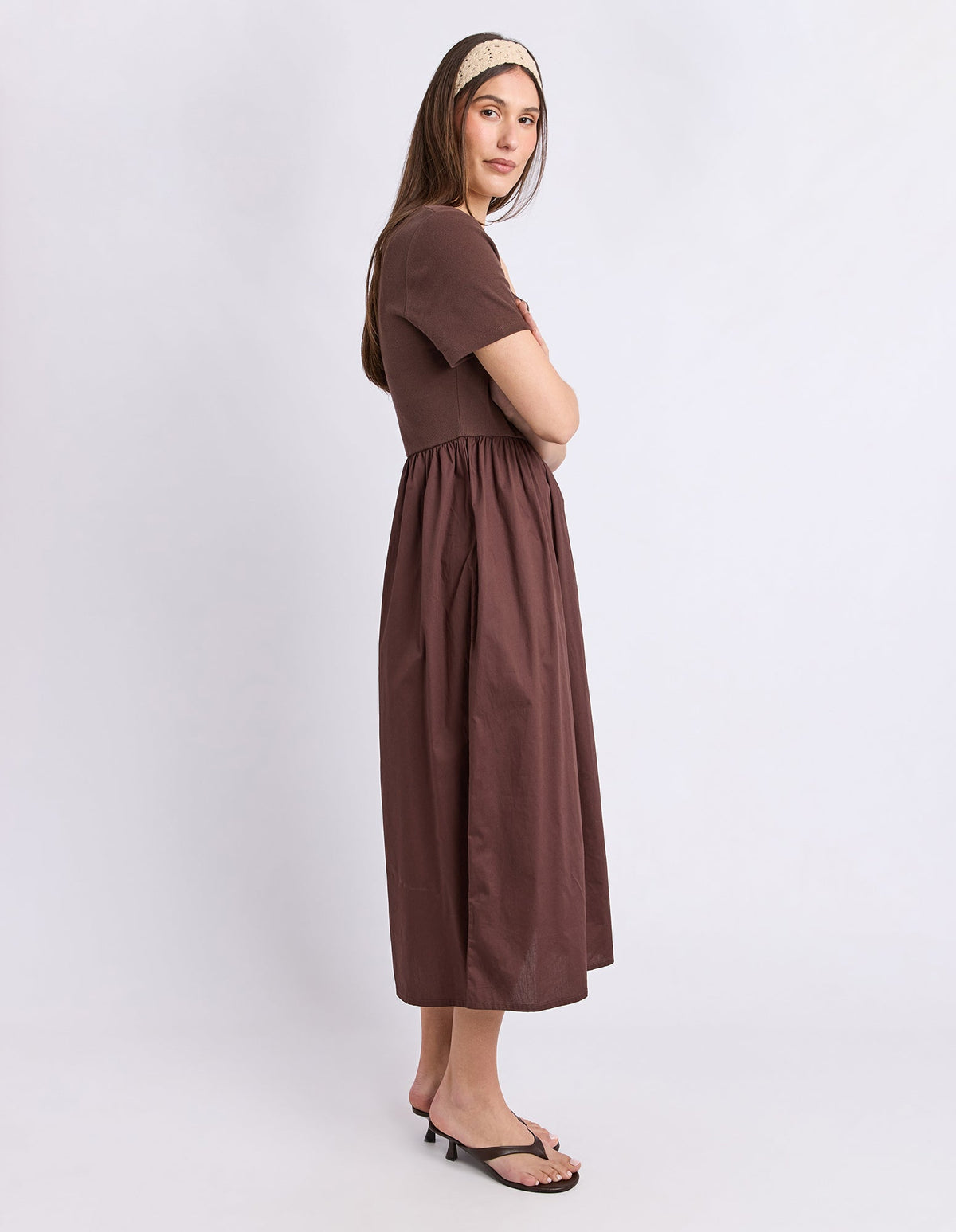 A woman modeling a brown short-sleeve midi dress with a gathered waist, paired with black heeled sandals and a beige patterned headband, standing against a light gray studio background.