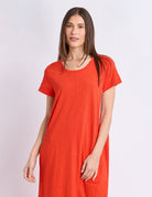 A woman modeling a short-sleeve, scoop-neck tunic dress in a vibrant orange-red color, set against a plain light gray studio background.