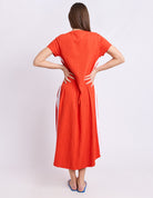 A woman viewed from the back wearing a bright orange short-sleeve midi dress with white side panels and blue sandals, standing against a light gray studio background.