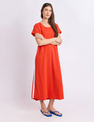 A woman modeling a bright orange-red linen midi dress with short flutter sleeves, a round neckline, and a white stripe detail along the side, paired with blue flip-flop sandals against a light gray studio background.