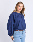Woman wearing a blue blouse with embroidery and jeans on a light gray background