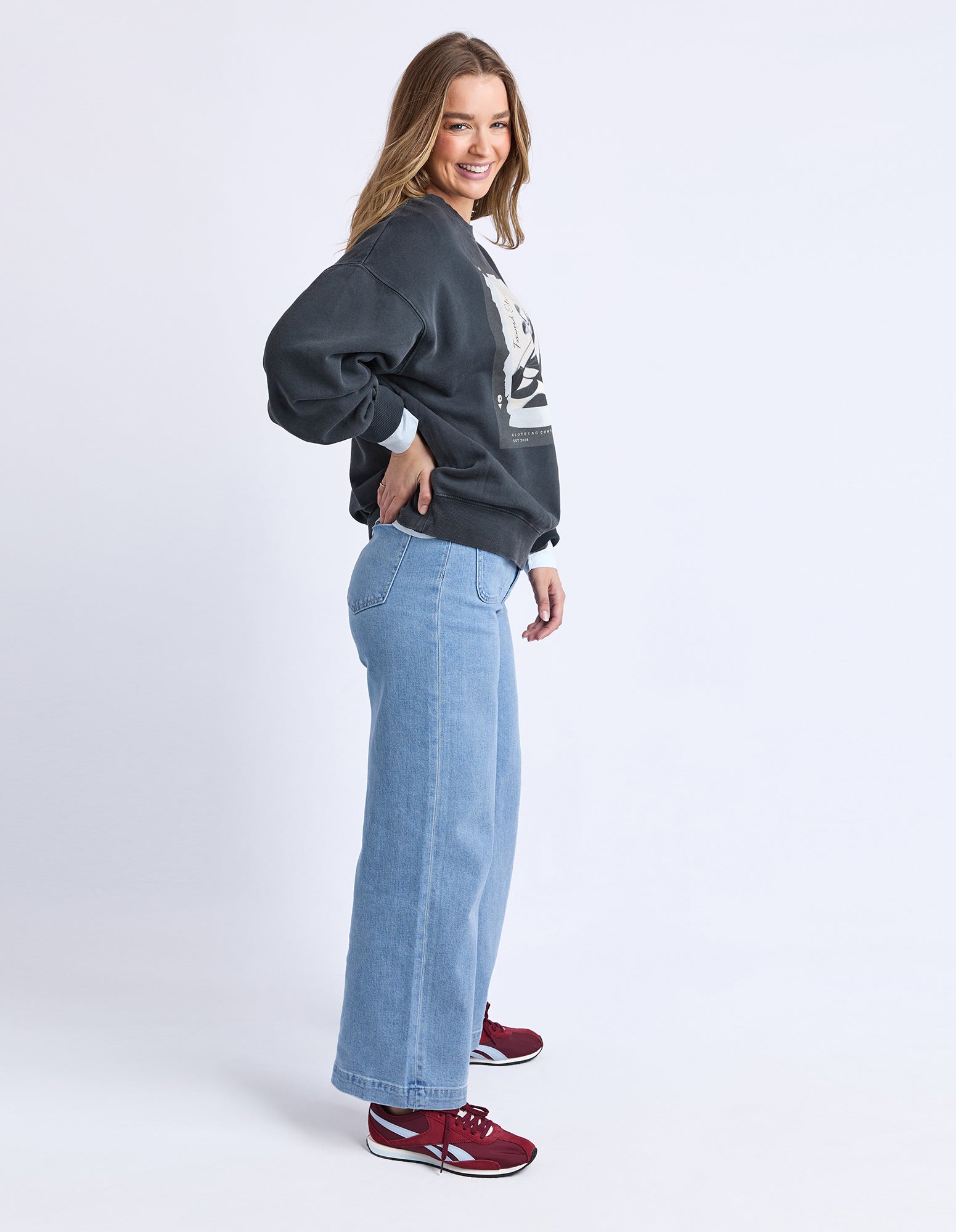 A woman modeling a dark charcoal gray sweatshirt, wide-leg light blue jeans, and burgundy sneakers against a plain white studio background.
