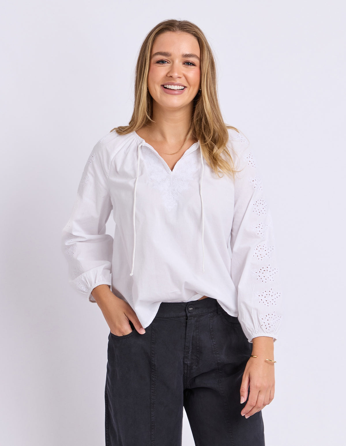 A woman wearing a white long-sleeve blouse with eyelet embroidery on the sleeves and a tie neckline, paired with dark charcoal pants, standing against a light gray studio background.