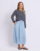 A woman modeling a navy and white horizontally striped long-sleeve top paired with a light blue midi skirt and black sandals, standing against a plain light gray studio background.