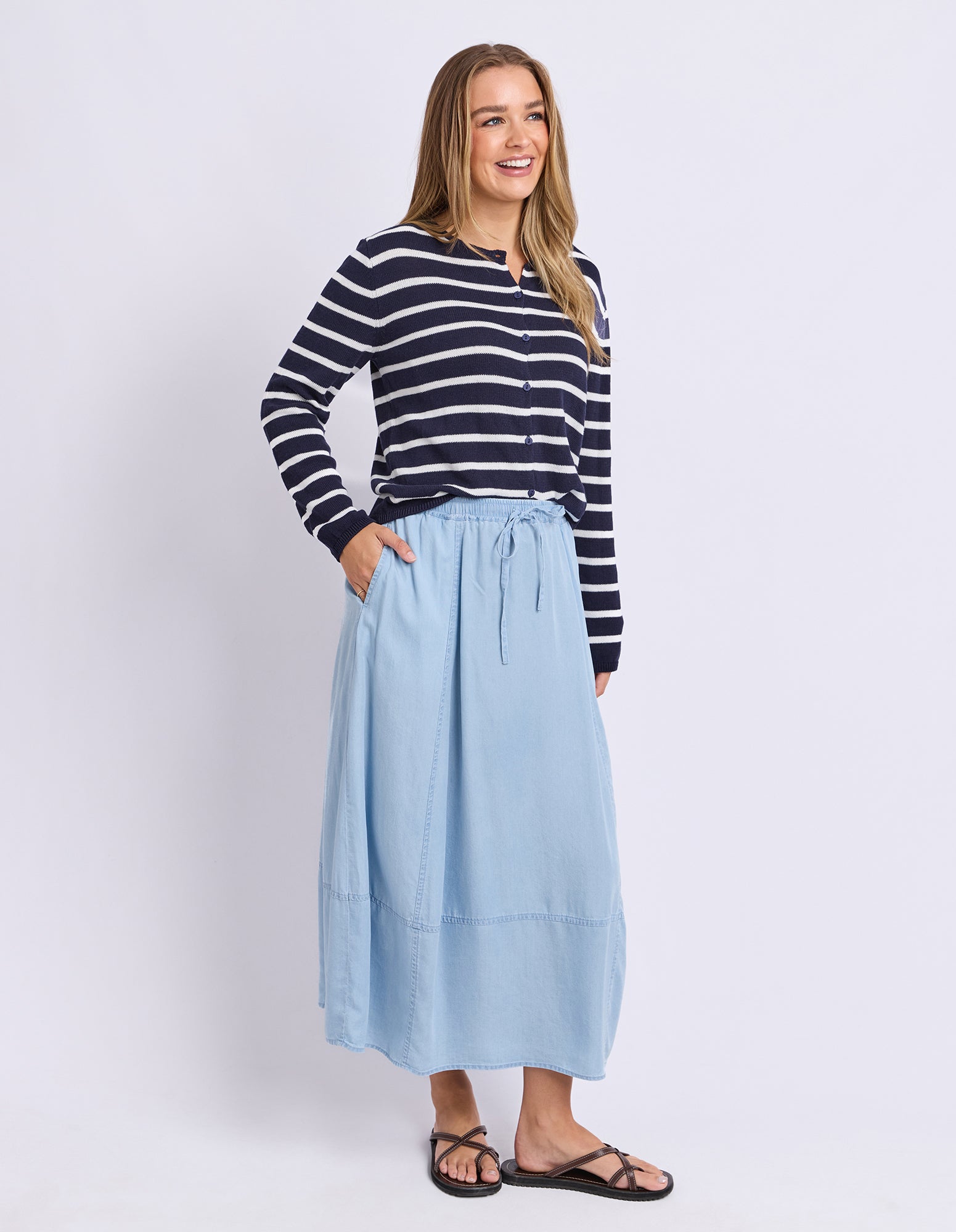 A woman modeling a navy and white horizontally striped long-sleeve top paired with a light blue midi skirt and black sandals, standing against a plain light gray studio background.