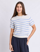 A woman modeling a short-sleeve knit sweater with thin horizontal light blue stripes on an off-white base, paired with dark navy pants, against a light gray studio background.