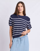 A woman modeling a navy blue short-sleeve knit top with white horizontal stripes, paired with light blue drawstring pants, against a light gray studio background.