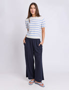 A woman modeling a short-sleeve knit top with light blue and white horizontal stripes, paired with dark navy wide-leg pants and light blue flip-flop sandals, against a plain light gray studio background.