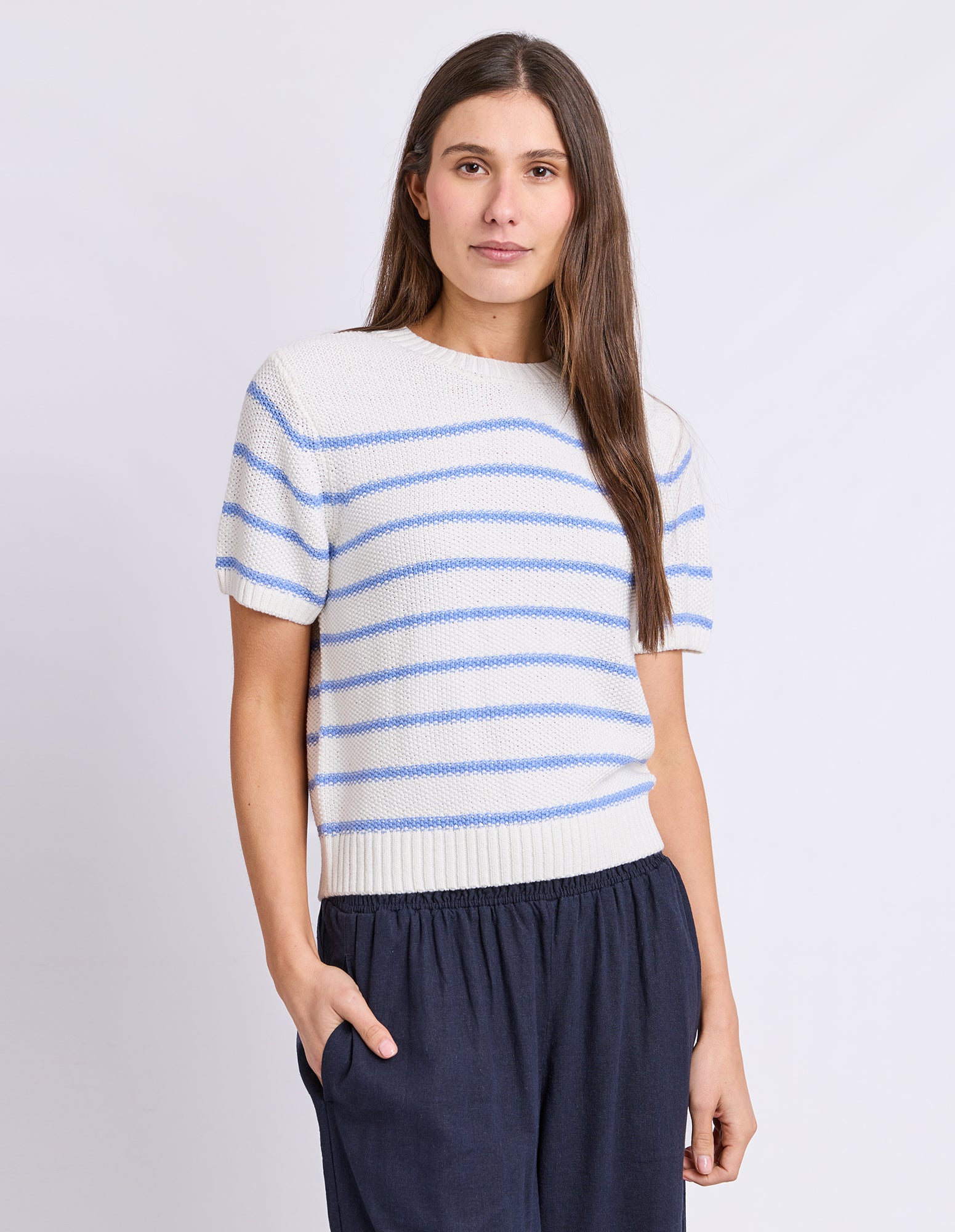 A woman modeling a short-sleeve knit top with horizontal light blue stripes on an off-white base, paired with dark navy pants, against a light gray studio background.