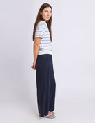 A woman modeling a short-sleeve knit top with light blue and white horizontal stripes, paired with dark navy wide-leg pants and blue sandals, photographed against a light gray studio background.