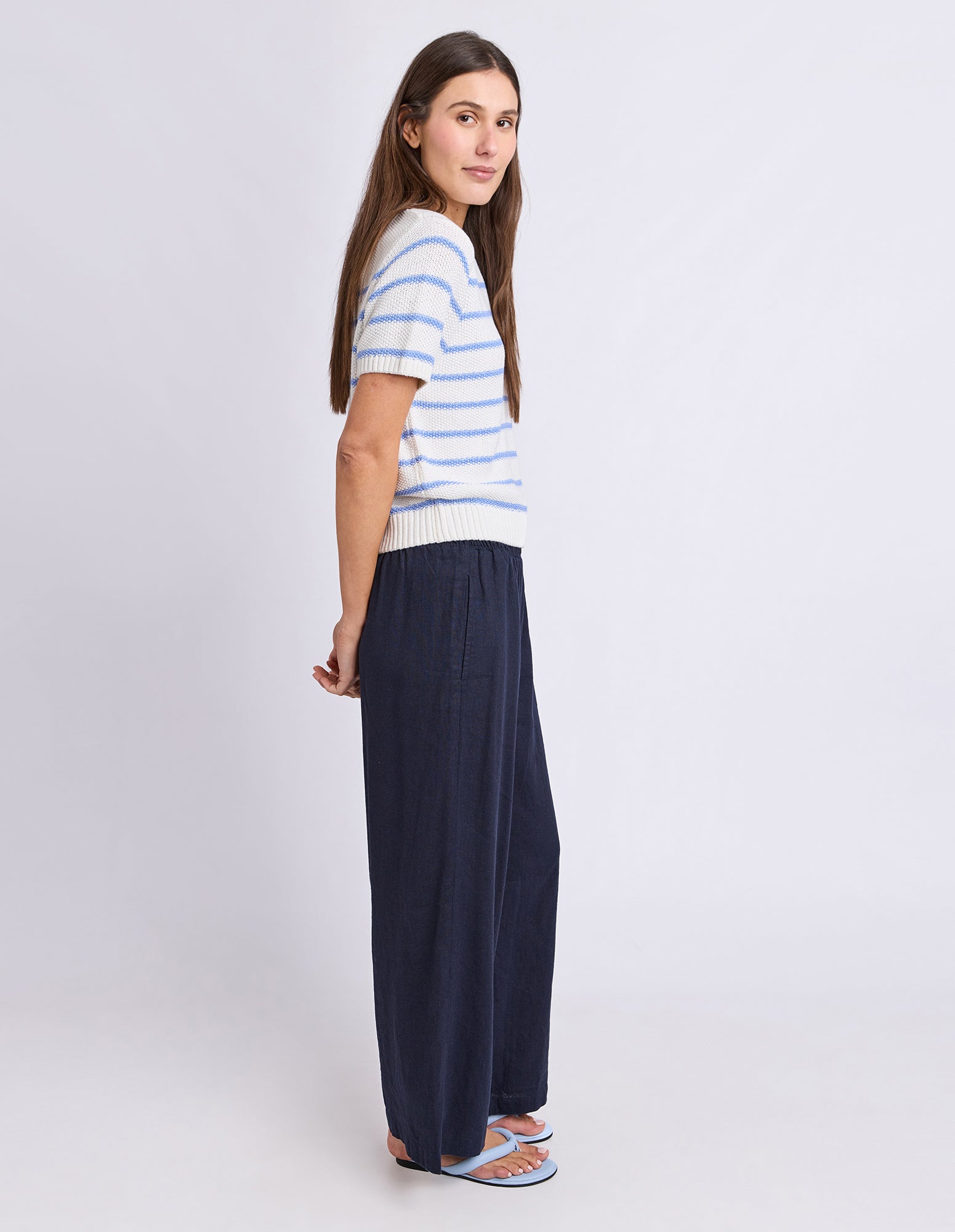 A woman modeling a short-sleeve knit top with light blue and white horizontal stripes, paired with dark navy wide-leg pants and blue sandals, photographed against a light gray studio background.