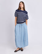 A woman modeling a navy blue short-sleeve knit top with white horizontal stripes and a light blue midi skirt, paired with dark brown flats, against a light gray studio background.