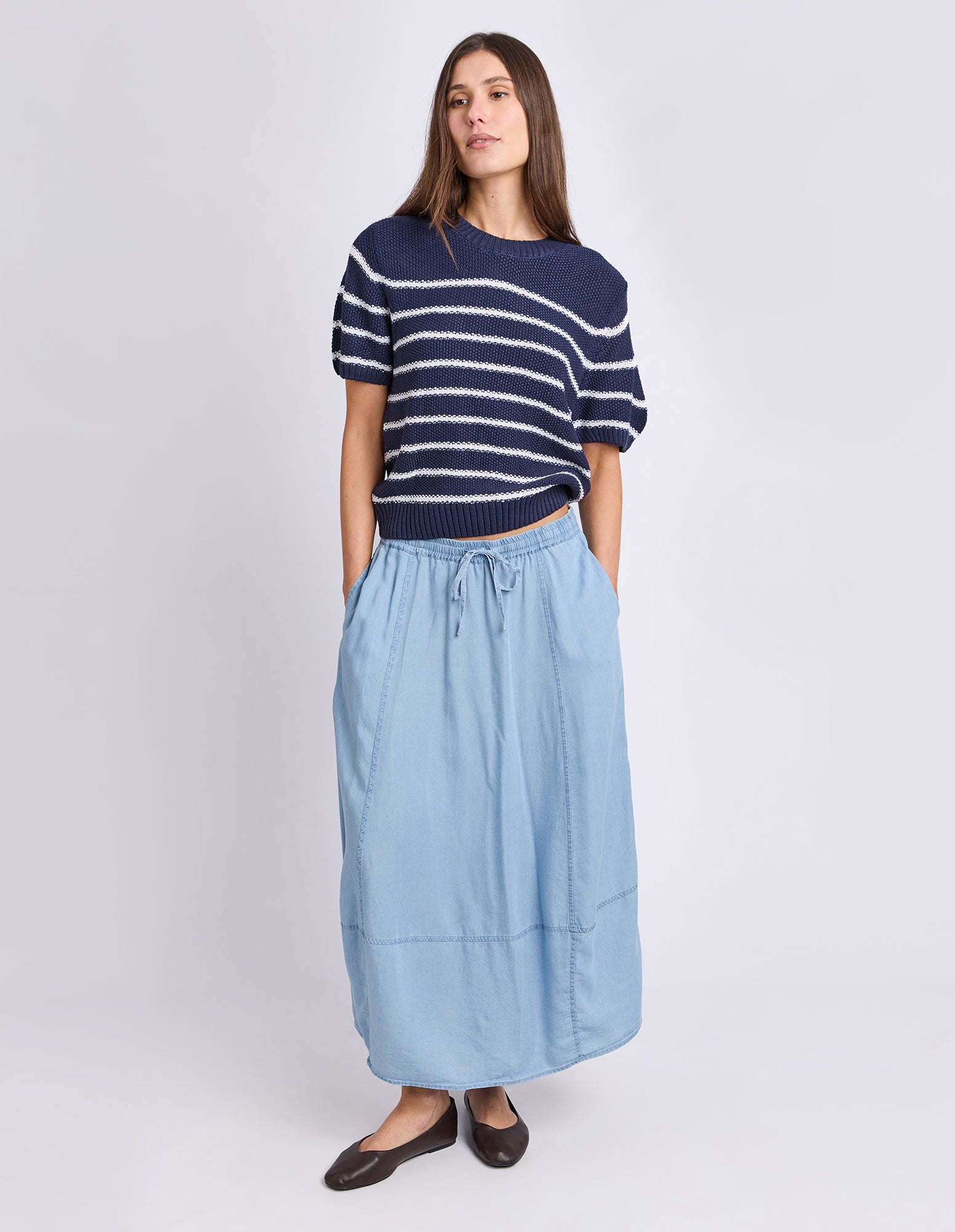 A woman modeling a navy blue short-sleeve knit top with white horizontal stripes and a light blue midi skirt, paired with dark brown flats, against a light gray studio background.