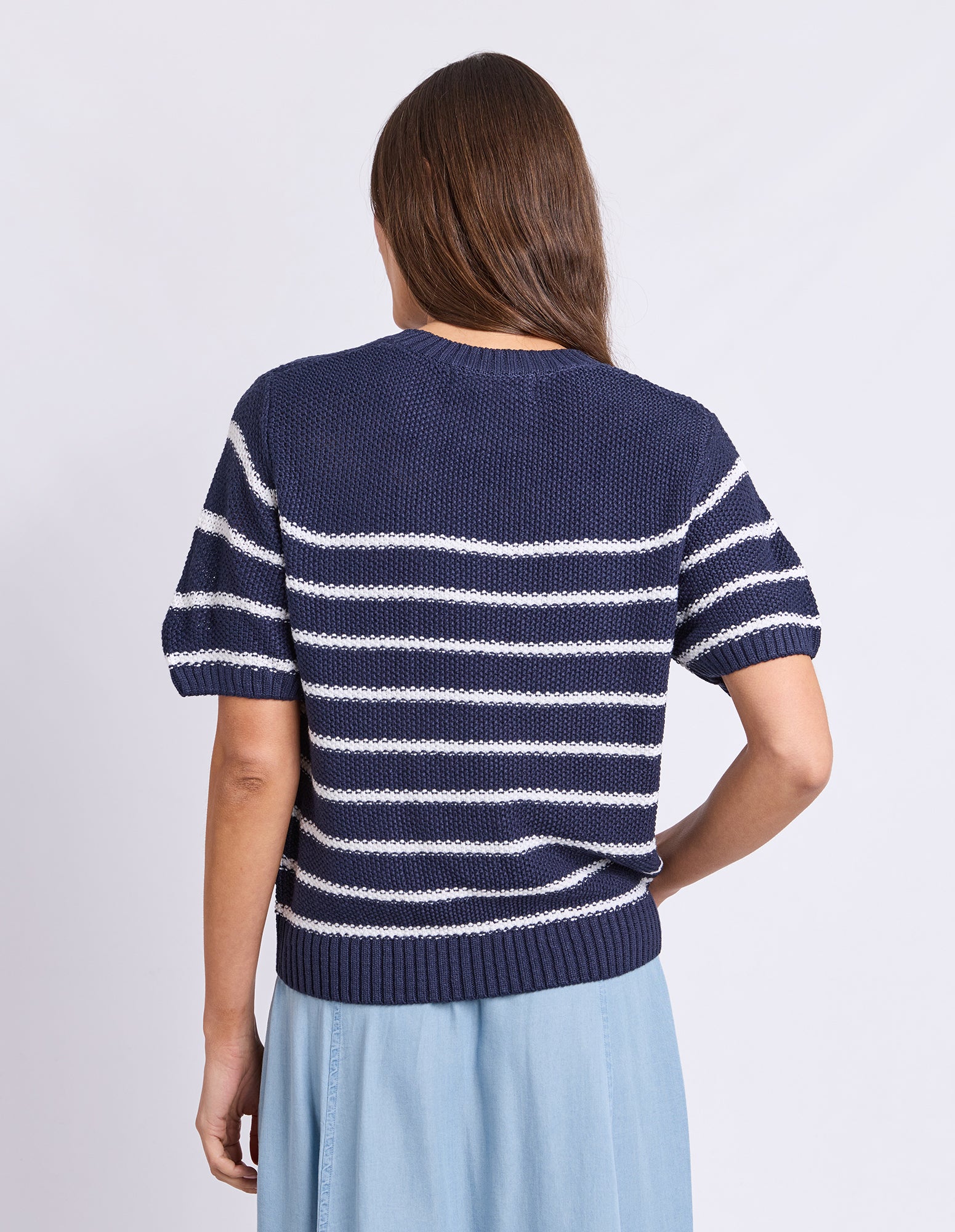 A woman viewed from the back wearing a navy blue short-sleeve knit sweater with white horizontal stripes and a light blue skirt, against a plain light gray studio background.