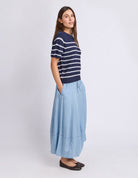 A woman modeling a short-sleeve knit top with navy and white horizontal stripes paired with a light blue maxi skirt and dark brown flats, set against a plain light gray studio background.