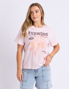 A woman modeling a light pink graphic t-shirt with 'FOXWOOD Media Design' printed on the front, paired with light blue denim shorts, against a plain light gray background.