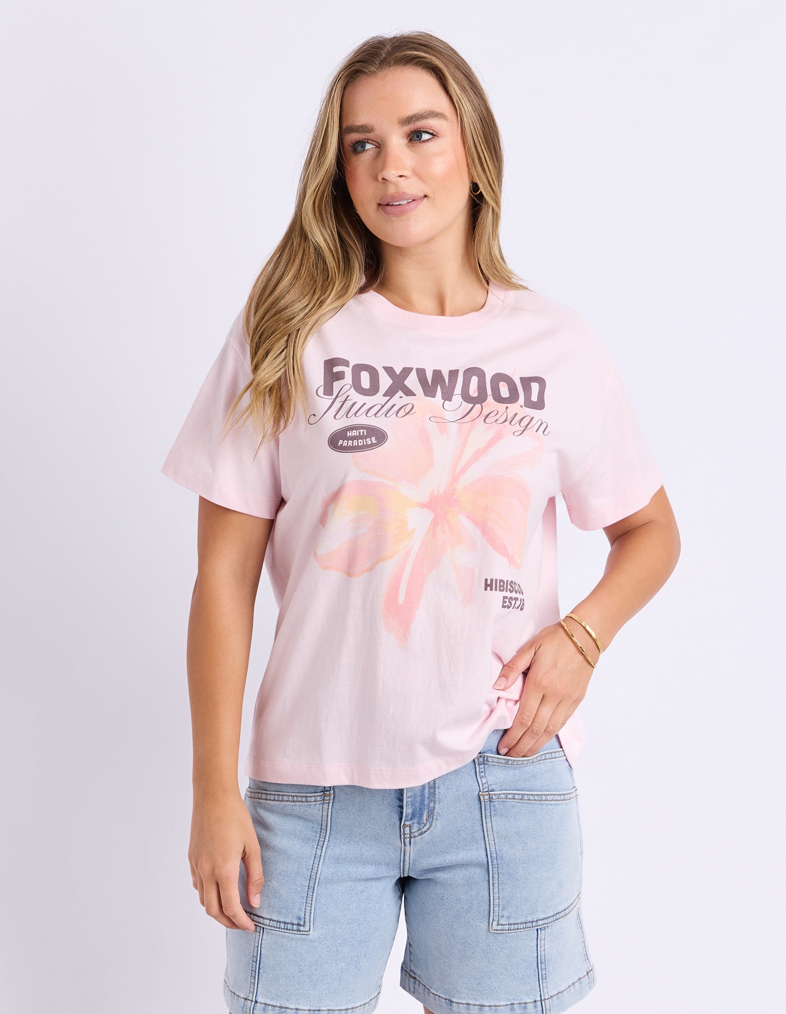 A woman modeling a light pink graphic t-shirt with 'FOXWOOD Media Design' printed on the front, paired with light blue denim shorts, against a plain light gray background.