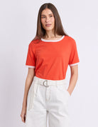 A woman modeling a bright orange short-sleeve ringer T-shirt with white contrast trim at the crew neck and sleeve cuffs, paired with white high-waisted trousers, against a light gray studio background.