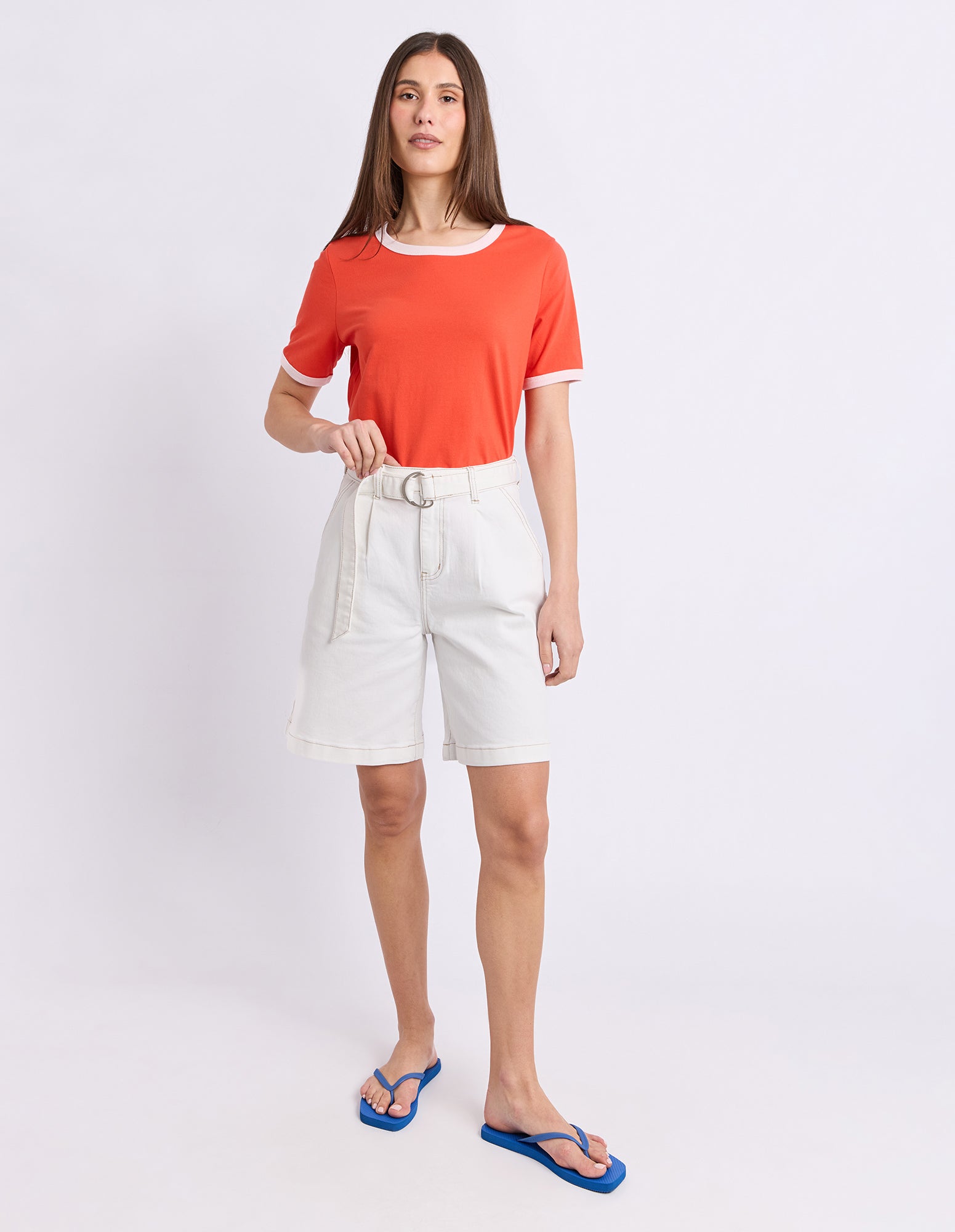 A woman modeling a bright orange short-sleeve ringer T-shirt with white trim, paired with white shorts, blue flip-flops, and a silver belt, set against a plain white studio background.