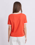 A woman viewed from the back wearing a bright orange short-sleeve ringer T-shirt with white trim at the collar and sleeve cuffs, paired with white pants, against a light gray studio background.